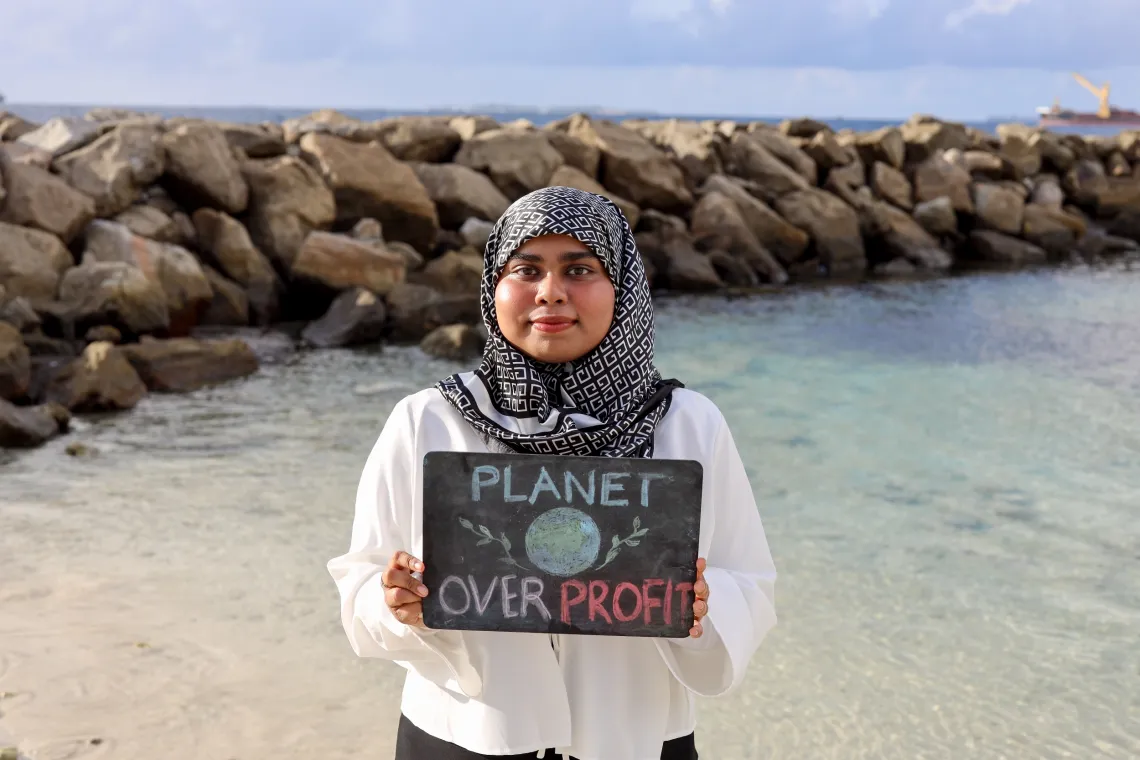a young woman stands on the ocean shore with a sign that reads Planet over profit