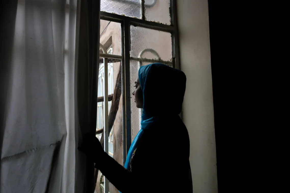 a woman in a hijab stands at a window looking out
