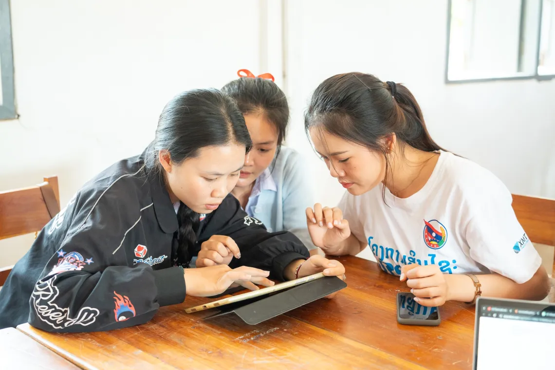 3 adolescent girls sit at a table around a tablet working on a project