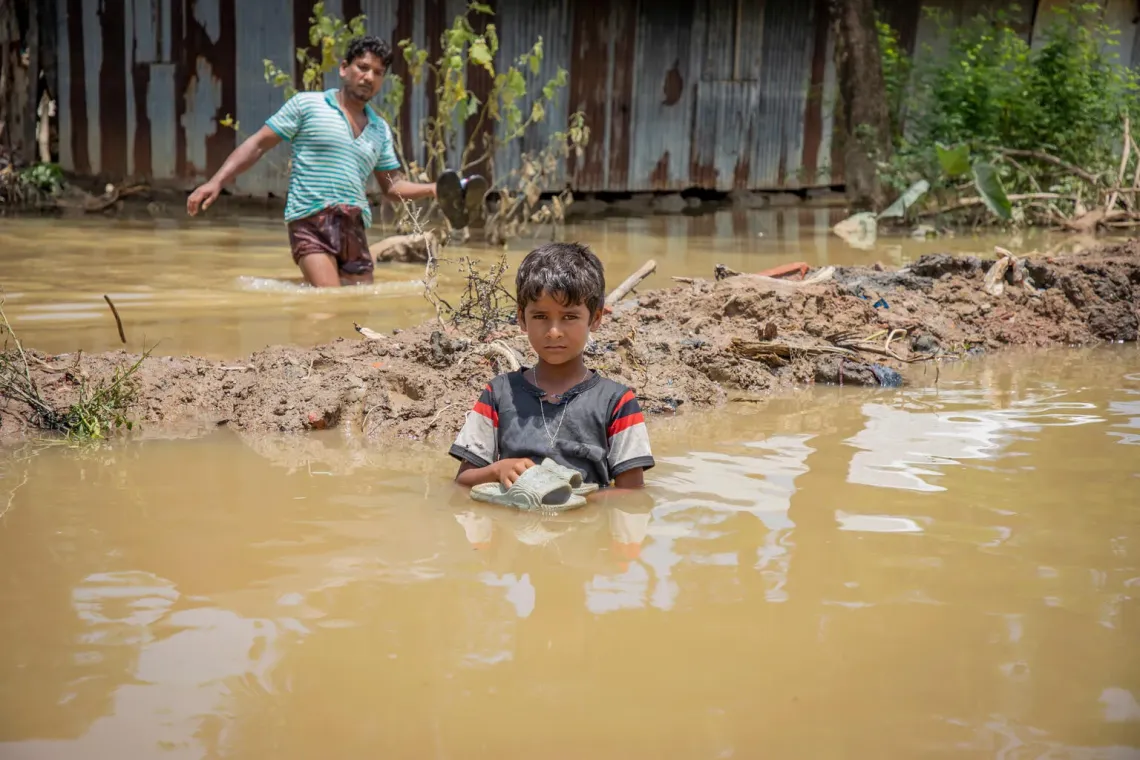 A boy stands waist deep in floodwaters holding his slippers.
