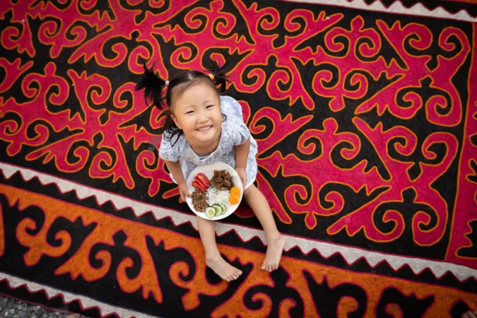 Nurayim, 3 years old, holds a plate of nutritious food at her home in Kurshab village, Uzgen district, Osh Oblast, Kyrgyzstan. Her mother tells us that she adapted well to eating food that the rest of the family eats &ndash; dairy products, meat fish, poultry, eggs, fruits and vegetables. The family avoids unhealthy foods in every way possible. They have not experienced any difficulties in obtaining wholesome foods in their region, and also have a garden where they grow some vegetables.