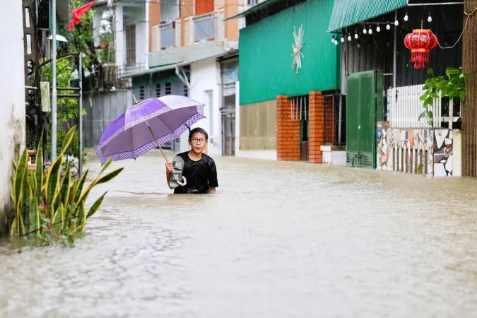 A boy wades through a flooded residential area, holding an umbrella and carrying their shoes.