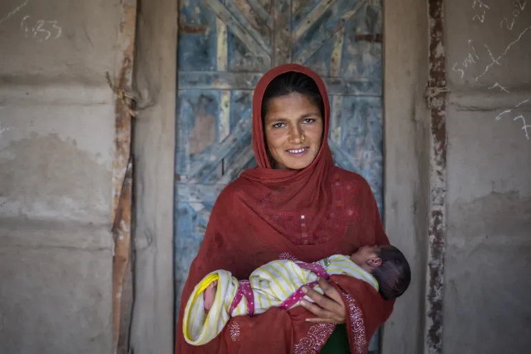 A woman smiles at the camera while gently holding a sleeping baby in her arms.