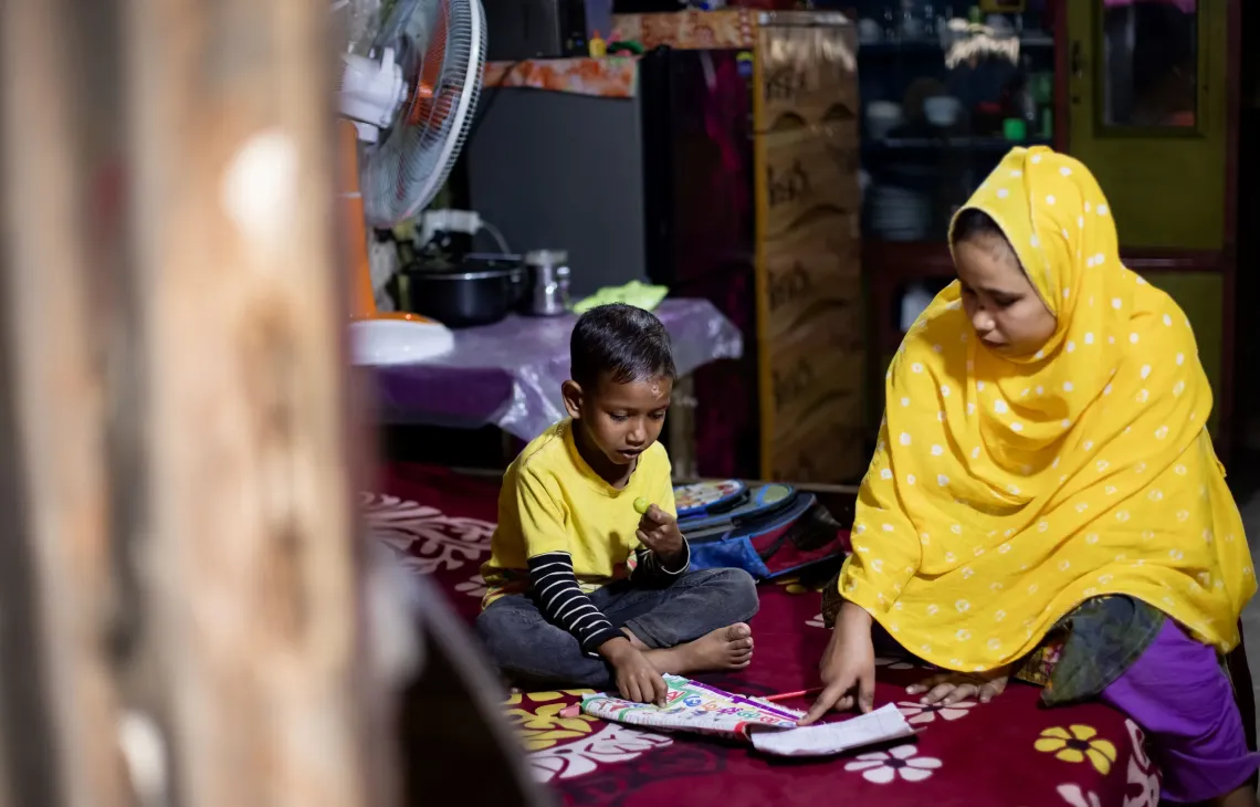 In their home, Pinky Akhter sits beside Muhammad as he goes through his lessons, trying to keep his education on track despite uncertainty.