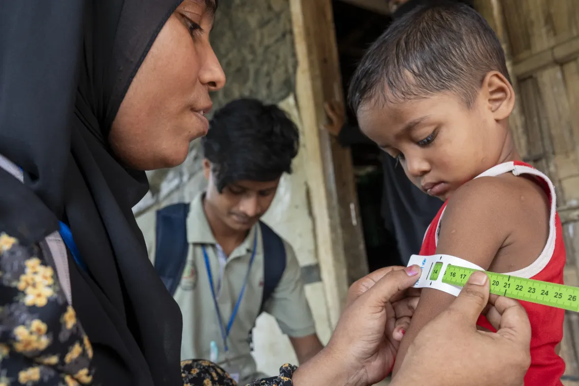 Tofura measures the Mid-Upper Arm Circumference (MUAC) of 31-month-old Shahed while Amin documents the measurements during a routine nutrition screening. 