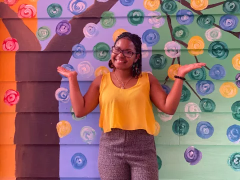 Belize: a young woman, Renata, stands in front of a colourful wall and raises her hands in a playful pose with a smile.