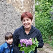 Žena s crvenkastom kosom drži buket jorgovana; djevojčica stoji pored, u vrtu kraj kamenog zida.