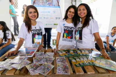 Tr&ecirc;s adolescentes participantes de projeto do UNICEF no Semi&aacute;rido est&atilde;o em p&eacute; atr&aacute;s de uma mesa com publica&ccedil;&otilde;es e folhetos