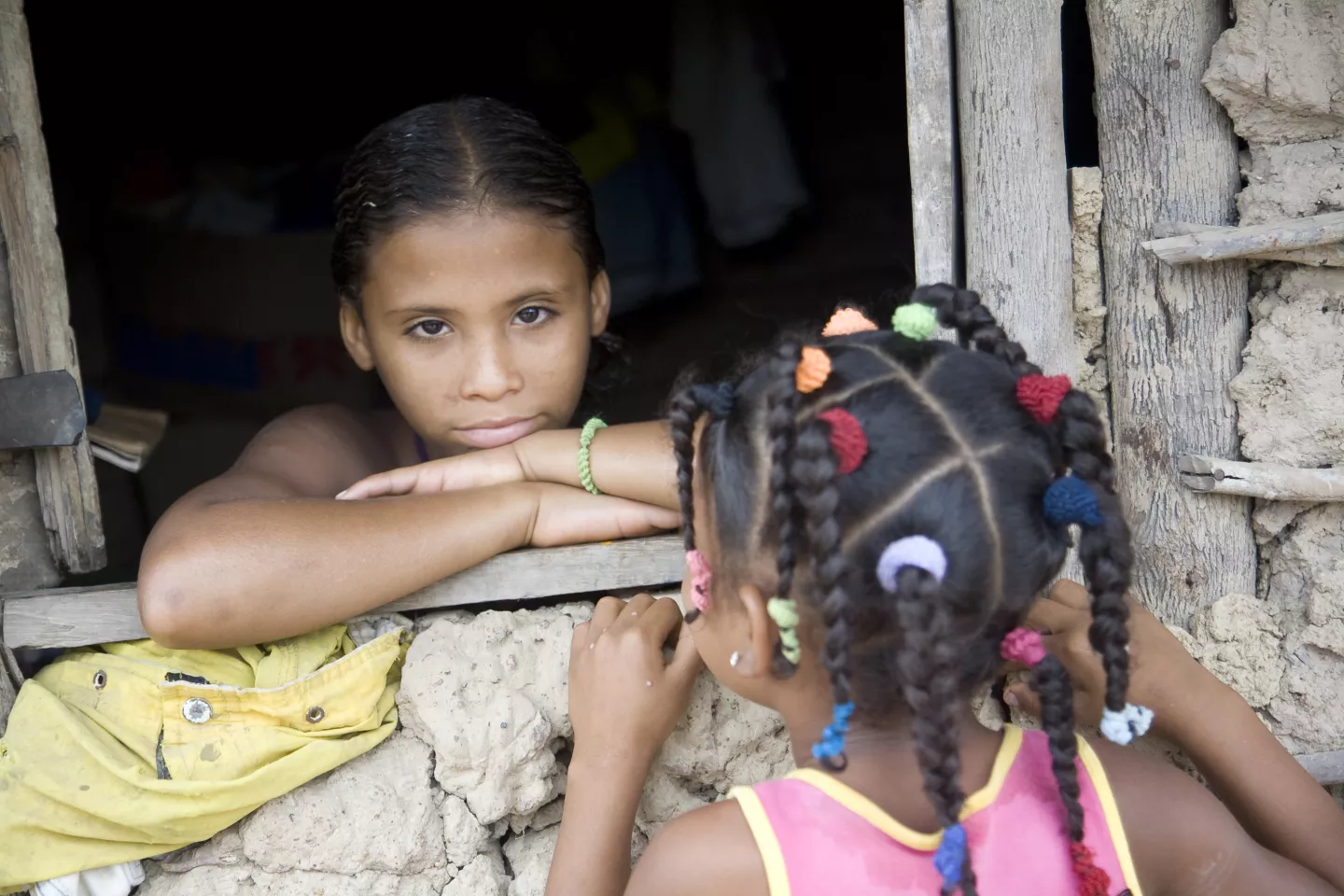 duas meninas est&atilde;o em uma janela de uma casa. uma do lado de dentro e a outra do lado de fora. a que est&aacute; do lado de dentro olha para a c&acirc;mera, a que est&aacute; do lado de fora est&aacute; de costas para a c&acirc;mera.