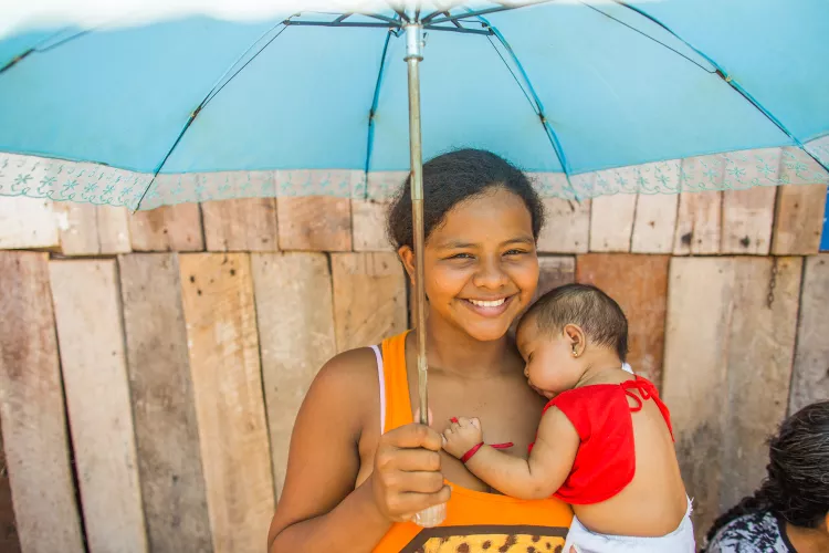 Mulher carrega sua filha beb&ecirc; no colo. Elas est&atilde;o embaixo de um guarda-chuva