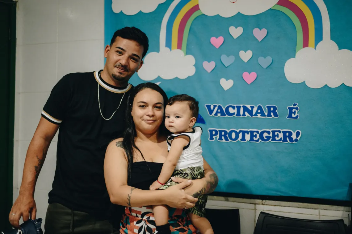 pai e m&atilde;e com filho no colo em frente a um painel sobre vacina&ccedil;&atilde;o 