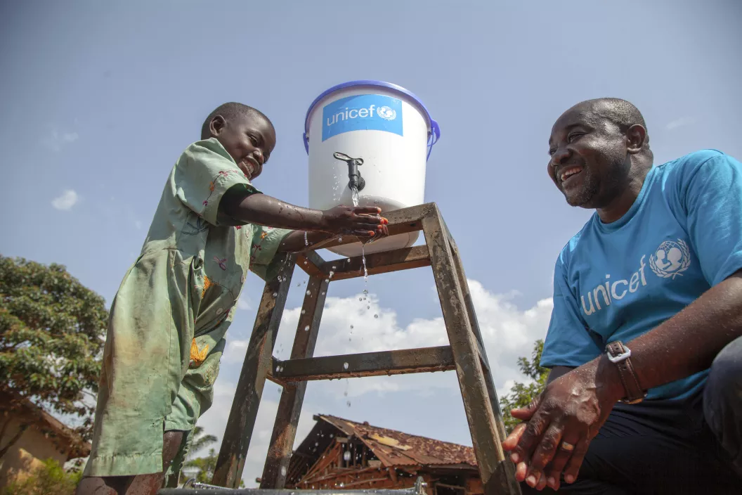 Na Rep&uacute;blica Democr&aacute;tica do Congo, um menino pequeno lava as m&atilde;os em um torneira que sai de um filtro com o logo do UNICEF. Ao lado dele, um funcion&aacute;rio do UNICEF usando camiseta azul com a marca da organiza&ccedil;&atilde;o. Os dois est&atilde;o em espa&ccedil;o aberto.