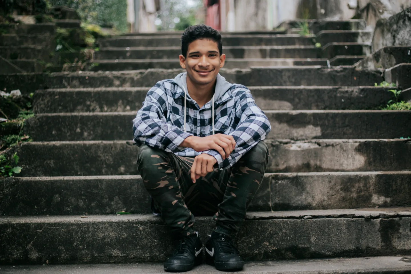 Foto mostra um adolescente sentado no degrau de uma escadaria. Ele est&aacute; olhando para a c&acirc;mera, sorrindo.