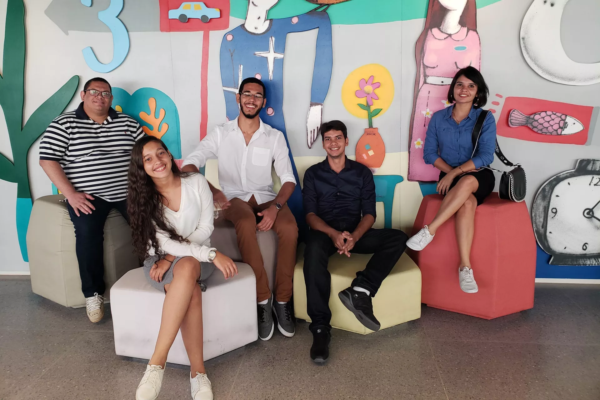 tr&ecirc;s jovens, uma adolescente e uma professora est&atilde;o sentados em pufes, olhando para a c&acirc;mera e sorrindo
