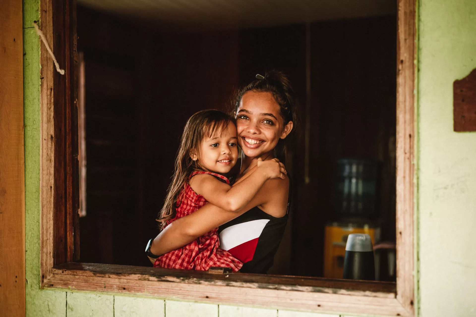 Foto mostra uma jovem segurando um menina pequena no colo. As duas est&atilde;o olhando para a foto e sorrindo.