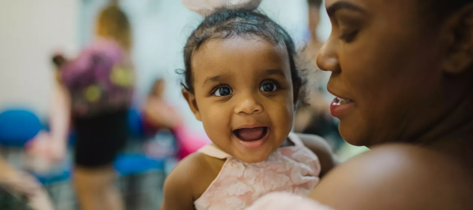 beb&ecirc; negra, de cabelos pretos presos, usando um la&ccedil;o cor de rosa na cabe&ccedil;a, veste camiseta rosa e &eacute; segurada no colo por uma mulher negra que aparece na lateral direita da foto 