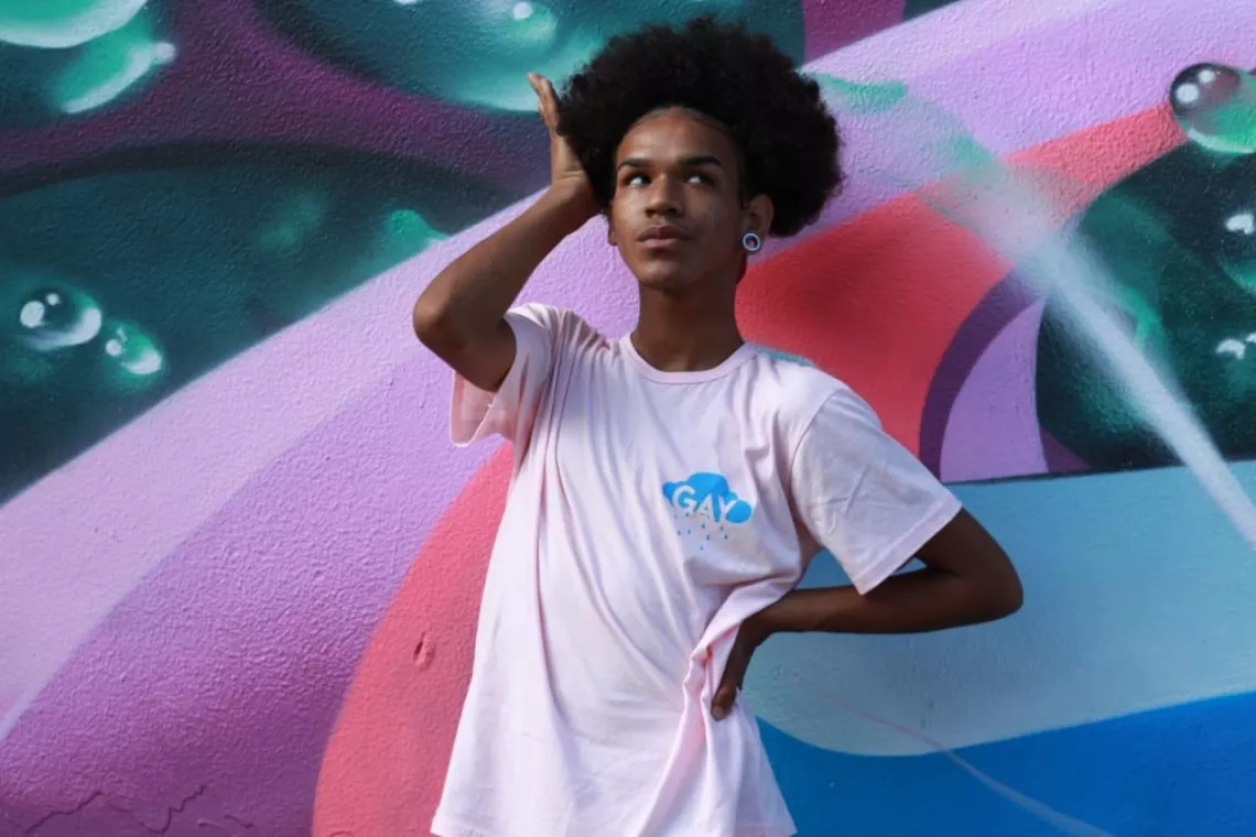 MC de 20 anos vence concurso de freestyle sobre o combate &agrave; Covid-19, promovido pelo Viva Melhor Sabendo Jovem. Na foto, o jovem est&aacute; parado em frente a um painel colorido, ele tem uma m&atilde;o na cabe&ccedil;a e outra na cintura.
