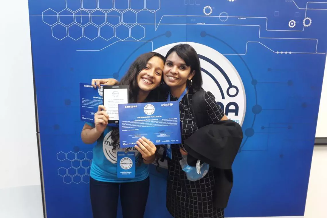 Uma adolescente e sua professora posam para a foto em frente a um painel da Maratona UNICEF Samsung. A adolescente segura um certificado nas m&atilde;os. As duas est&atilde;o sorrindo.