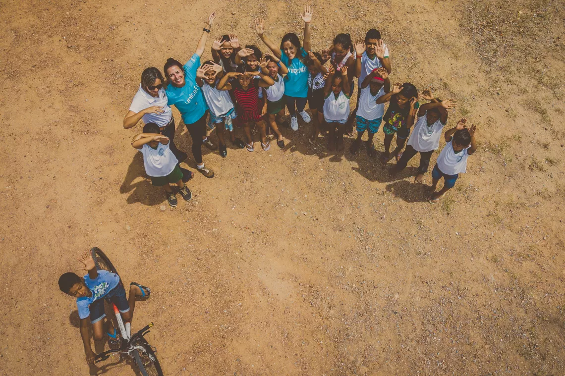 Foto a&eacute;rea de um grupo de pessoas. Elas est&atilde;o em p&eacute; em uma estrada de terra. H&aacute; alguns adultos e v&aacute;rias crian&ccedil;as. Alguns est&atilde;o com os bra&ccedil;os levantados. No meio do grupo, h&aacute; duas mulheres com a camiseta do UNICEF. No canto inferior esquerdo, aparece um menino de bicicleta.