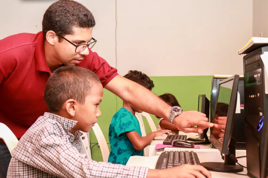 em uma sala de inform&aacute;tica, um professor, de p&eacute;, mostra algo na tela do computador, para um menino que est&aacute; sentado.