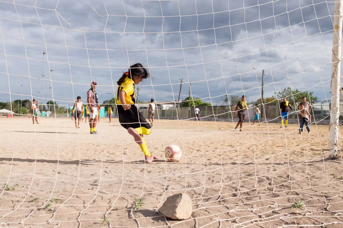 menina chuta bola de futebol. ela est&aacute; vestida com um uniforme amarelo e o campo &eacute; de terra