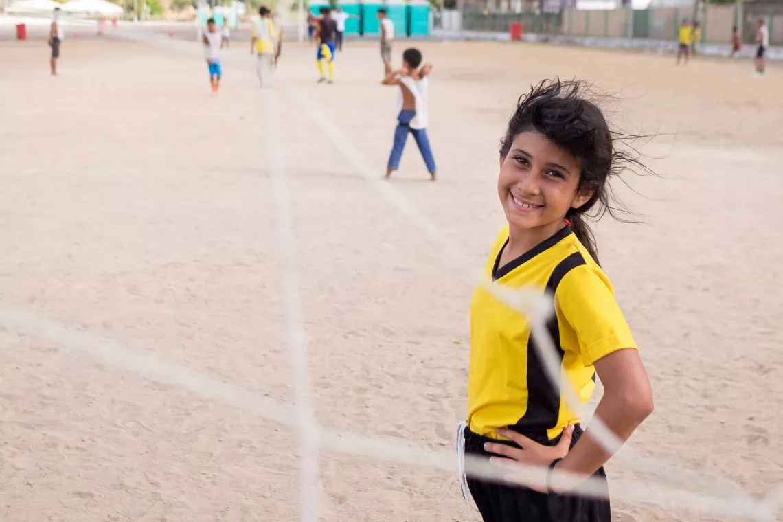 menina olha para a c&acirc;mera atrav&eacute;s da rede do gol. ela veste um uniforme amarelo. o campo &eacute; de terra.