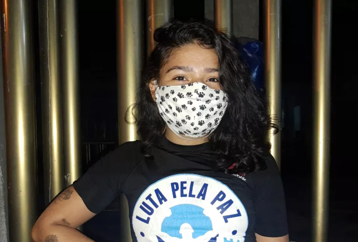 Lorena olha para a c&acirc;mera, sorrindo com os olhos. Ela usa m&aacute;scara e uma camiseta com o logo da Luta pela Paz