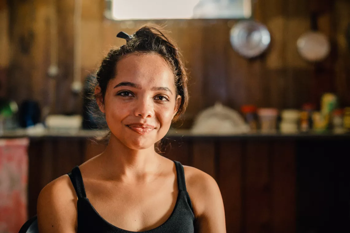 Foto mostra uma jovem sorrindo para a c&acirc;mera. Ela est&aacute; em um ambiente interno. O fundo da foto est&aacute; desfocado.