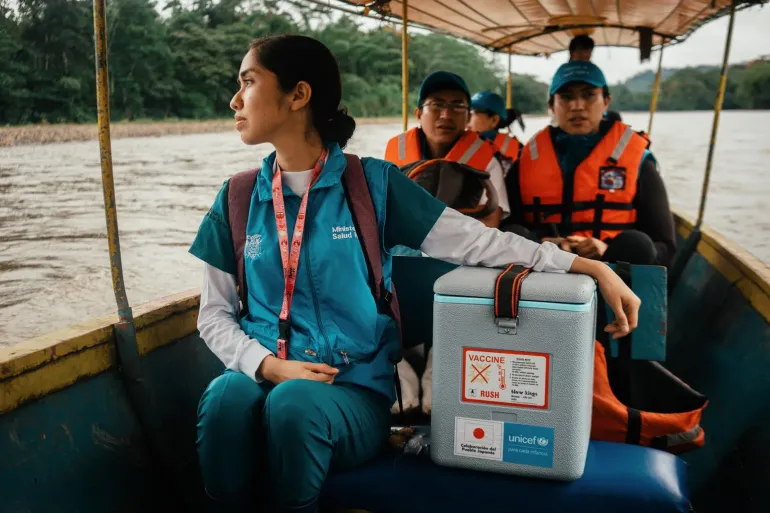 mulher vestida de uniforme azul de enfermeira segura caixa t&eacute;rmica de vacinas, em um barco com outras pessoas atr&aacute;s