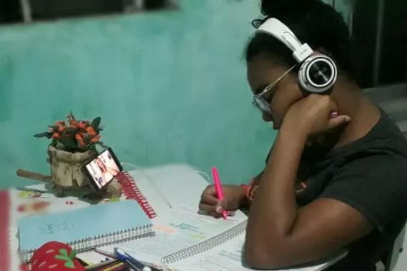 Dedicada aos estudos, a adolescente Luziana Flora descobriu no projeto Gera&ccedil;&atilde;o que Move uma oportunidade de aprender mais sobre seu territ&oacute;rio. Foto: adolescente, usando fone de ouvido, estuda sentada &agrave; mesa de sua casa. na mesa h&aacute; cadernos, livros e material escolar