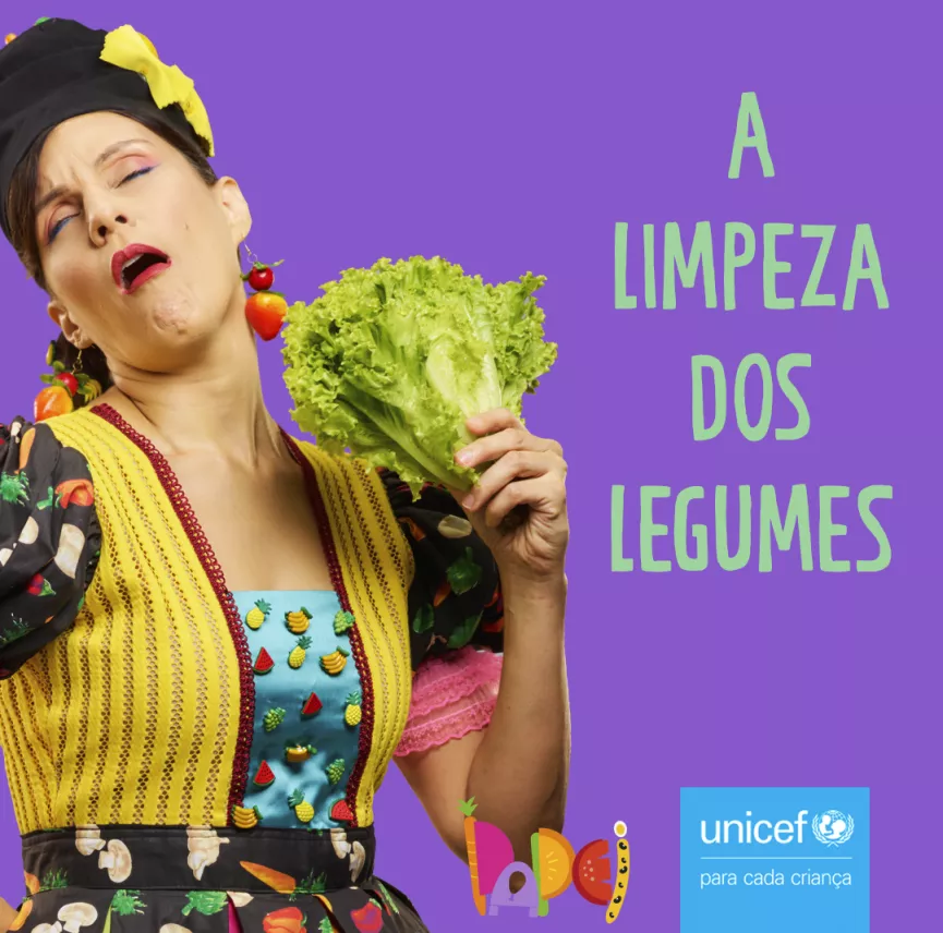 Card com fundo roxo, mostra uma mulher se abanando com um p&eacute; de alface. Est&aacute; escrito em verde: A limpeza dos legumes. No canto inferior direito aparecem os logos da campanha e do UNICEF.