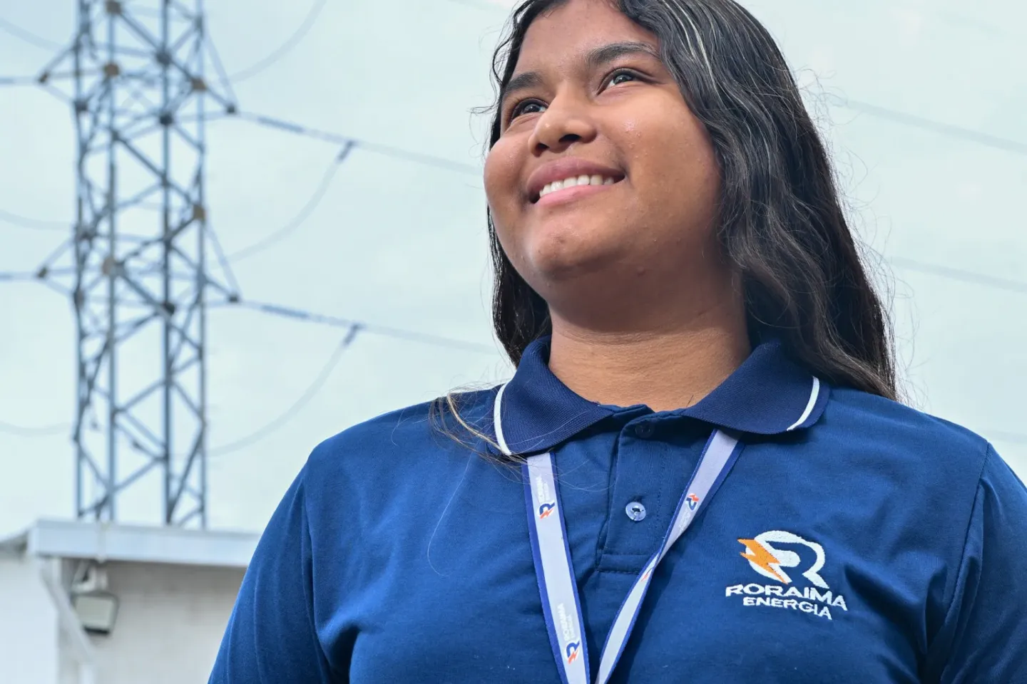 jovem de cabelos escuros vestindo uniforme de trabalho azul olha para o lado esquerdo, ao fundo se v&ecirc; uma torre de distribui&ccedil;&atilde;o de energia el&eacute;trica