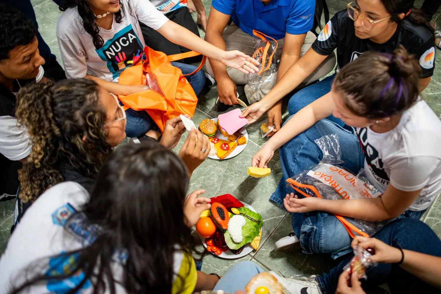 Foto mostra v&aacute;rios adolescentes sentados no ch&atilde;o em volta de pratos de comida