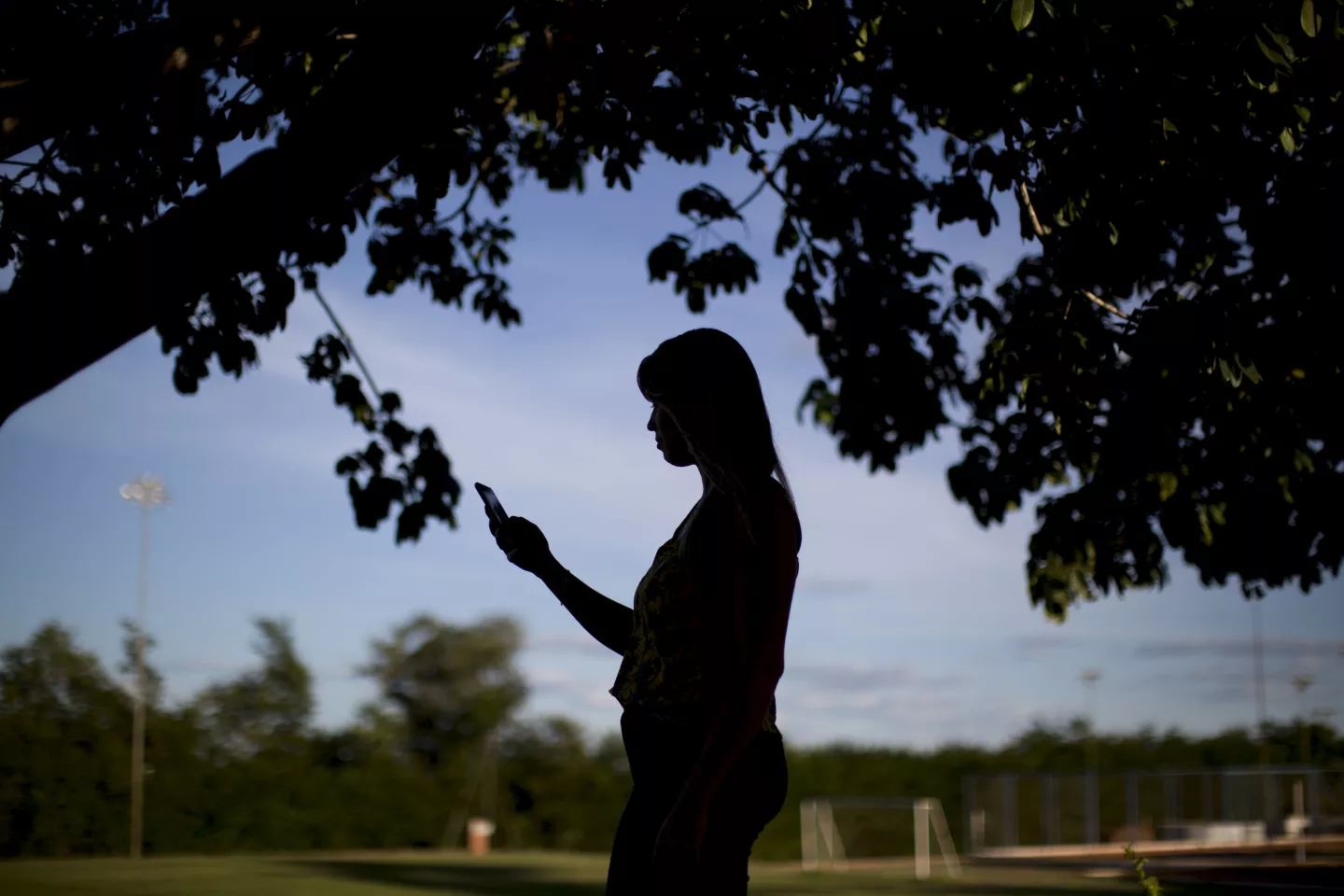 a foto mostra a silhueta de uma adolescente ao entardecer. ela est&aacute; de perfil e segura um celular.