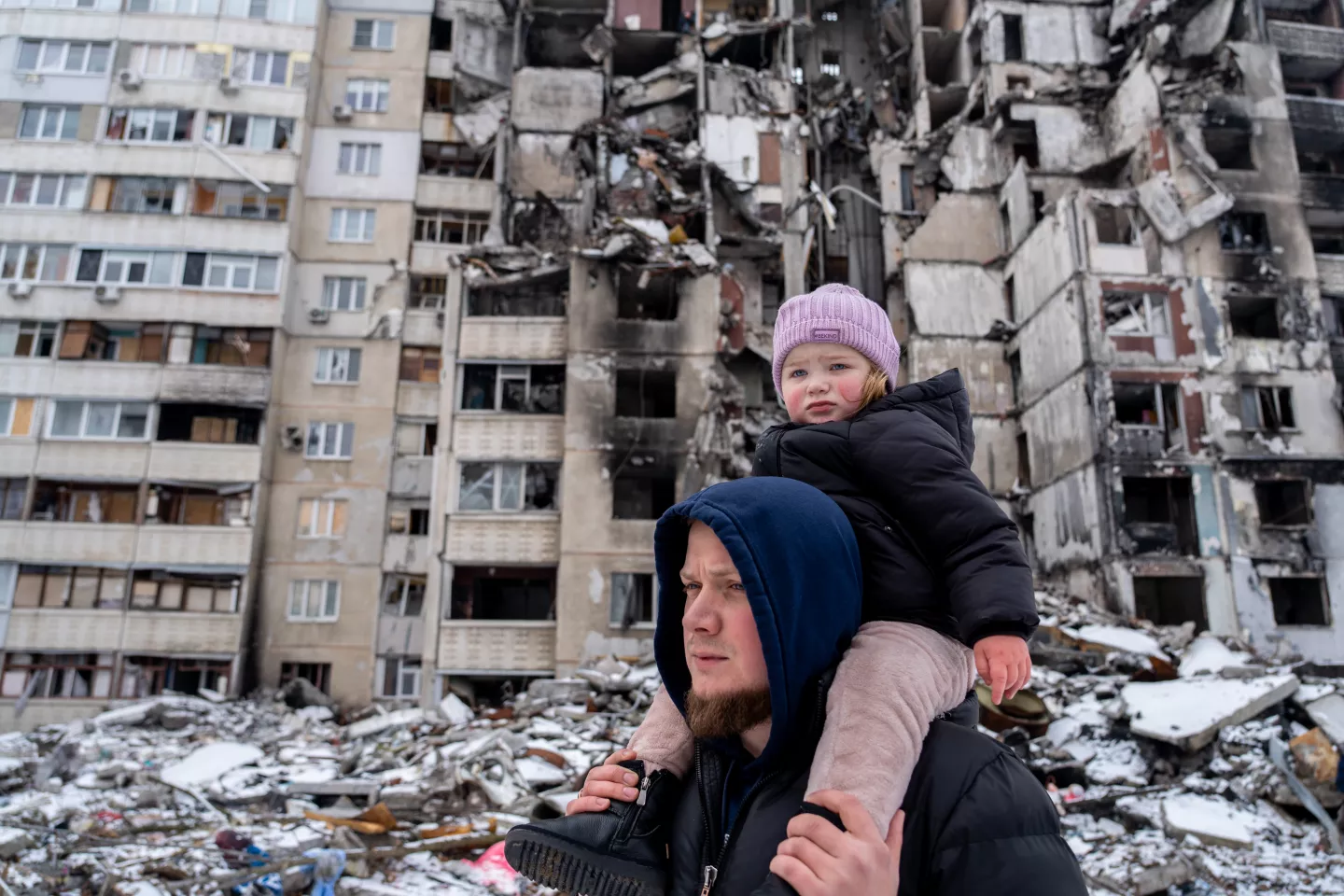 Foto mostra um homem carregando uma beb&ecirc; no ombro. Atr&aacute;s deles, h&aacute; pr&eacute;dios destru&iacute;dos pela guerra.