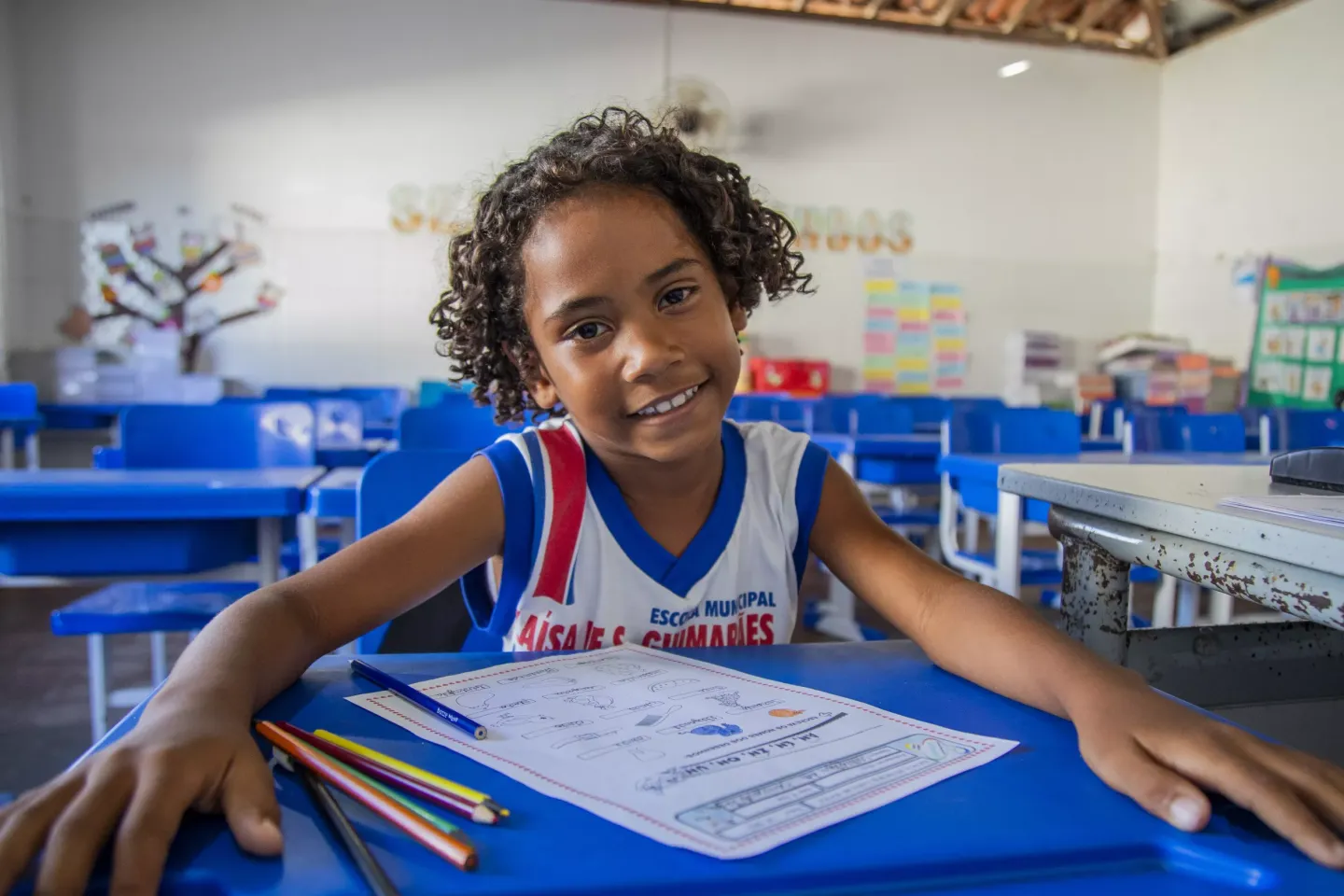 Foto mostra Julia sentada na mesa escolar, sorrindo.