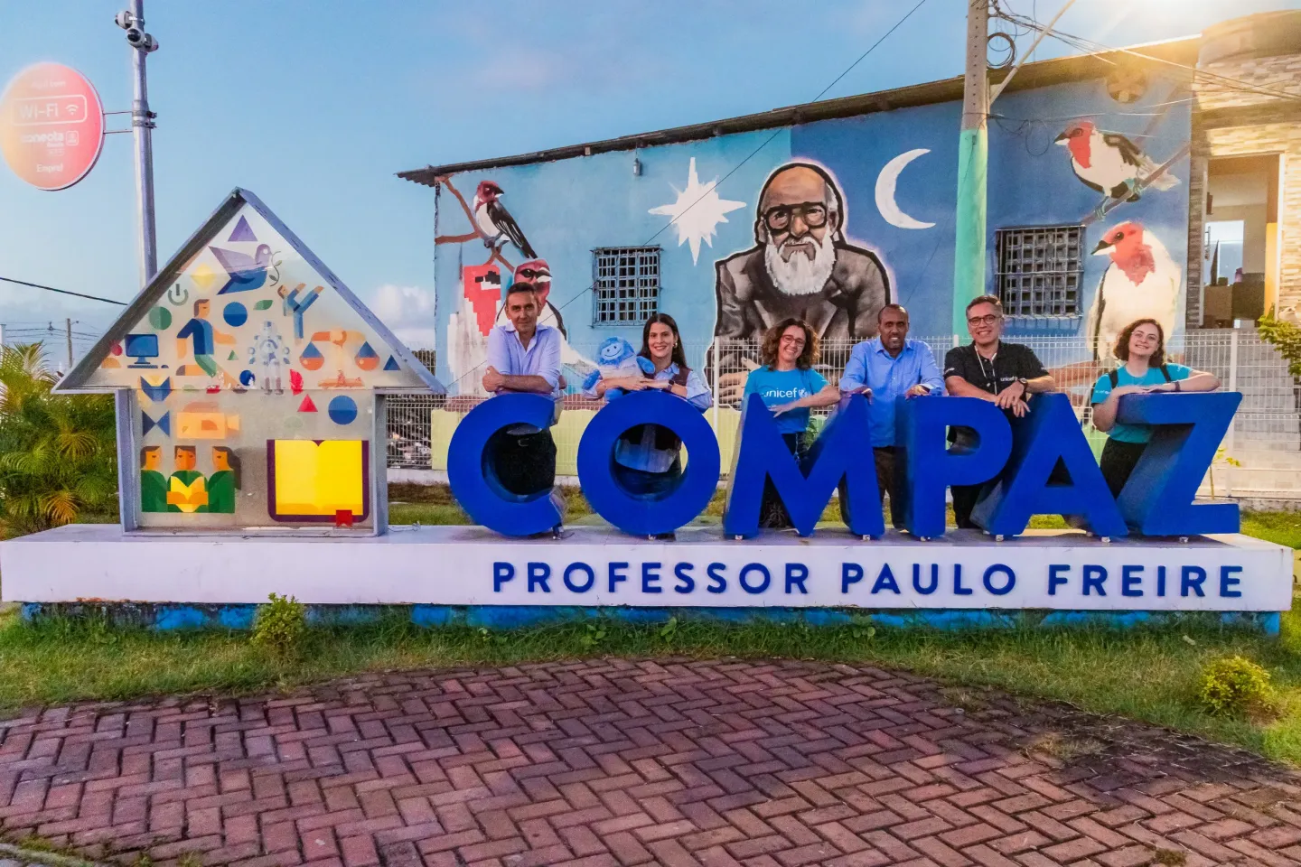 pessoas atr&aacute;s de uma placa onde se l&ecirc; COMPAZ