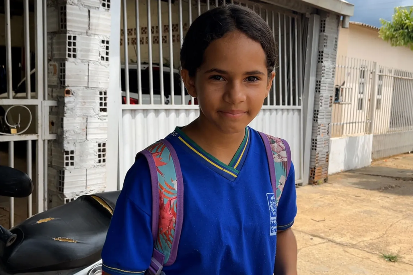 Foto mostra menina com uniforme escolar e mochila pendurada nas costas. Ela est&aacute; olhando para a c&acirc;mera.