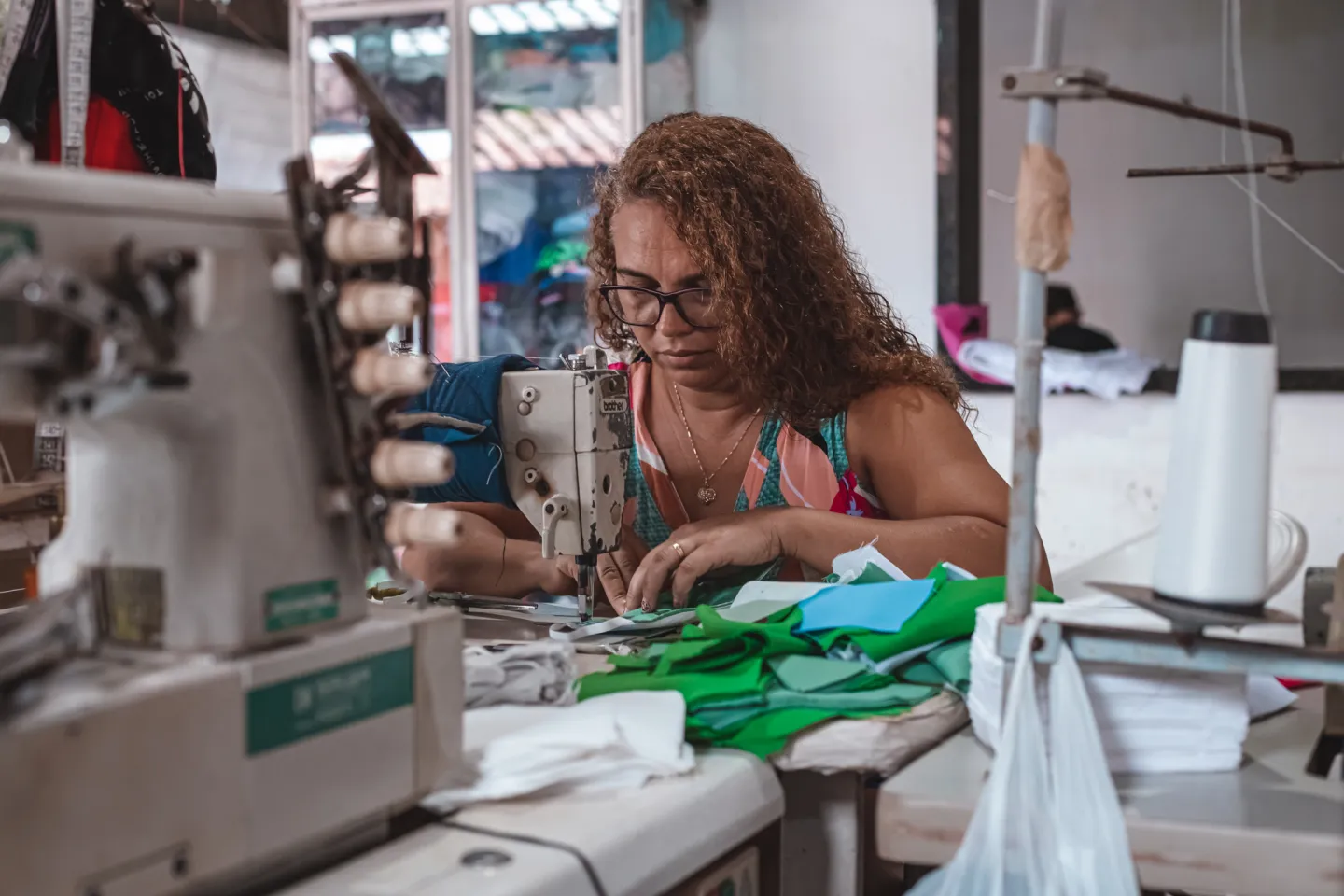 Foto mostra um mulher costurando em uma m&aacute;quina de costura. Em volta dela h&aacute; diversos materiais de costura, como linhas e tecidos.
