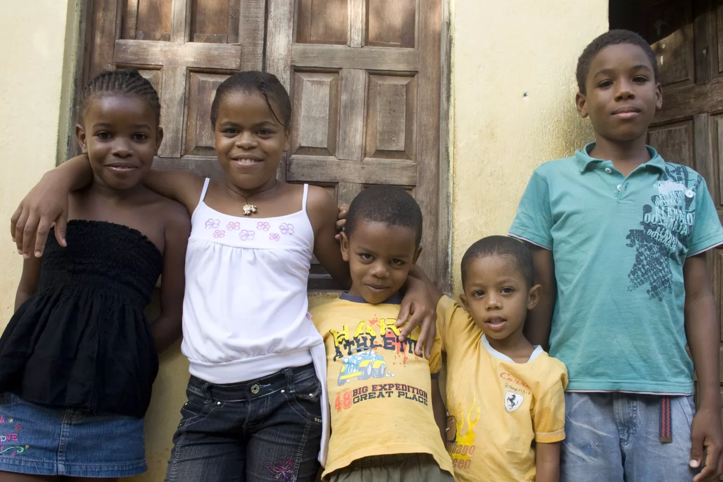 Cinco crian&ccedil;as de diferentes idades est&atilde;o encostadas em uma parede externa. Elas olham para a c&acirc;mera.