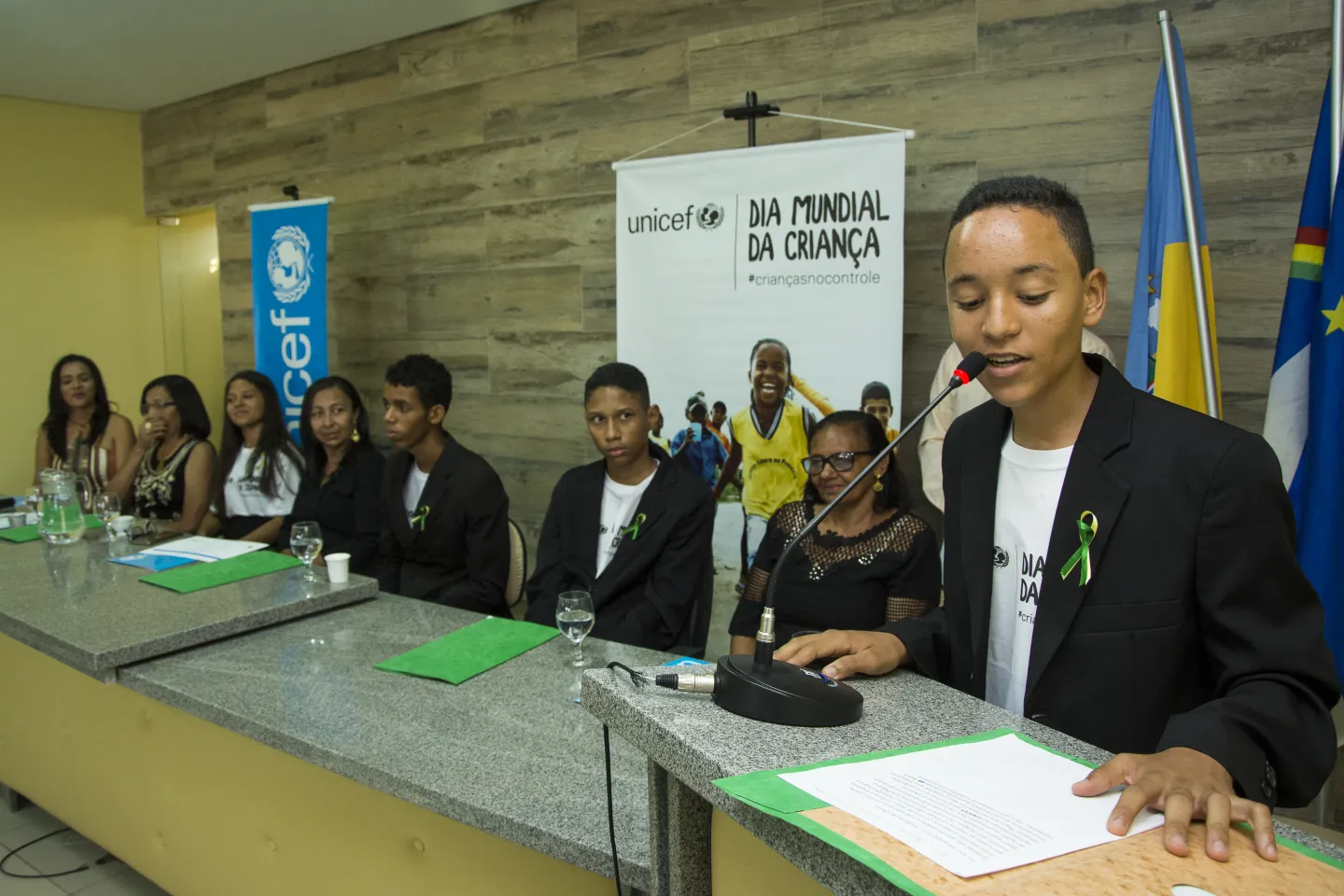 Adolescente fala em tribuna. Atr&aacute;s dele, em uma mesa, est&atilde;o outros adolescentes e um banner do Dia Mundial da Crian&ccedil;a