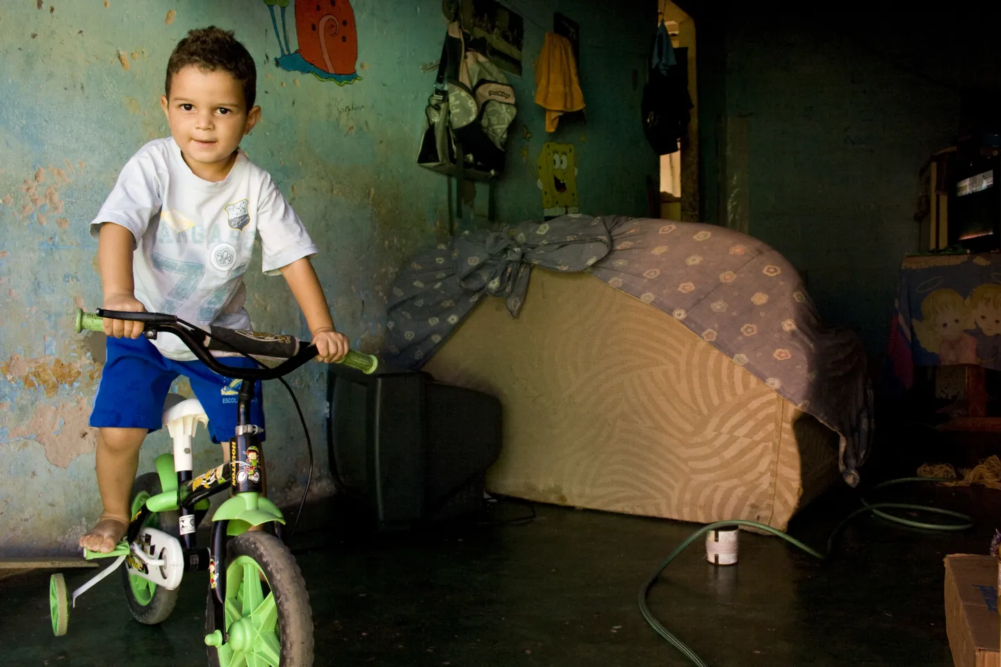 um menino pequeno est&aacute; em cima de uma bicicleta dentro da sala de casa, ao fundo vemos o sof&aacute;/