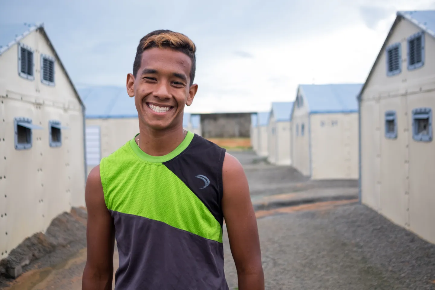 o adolescente Santiago est&aacute; na frente de um acampamento, ele usa uma camiseta sem mangas e sorri para a c&acirc;mera.