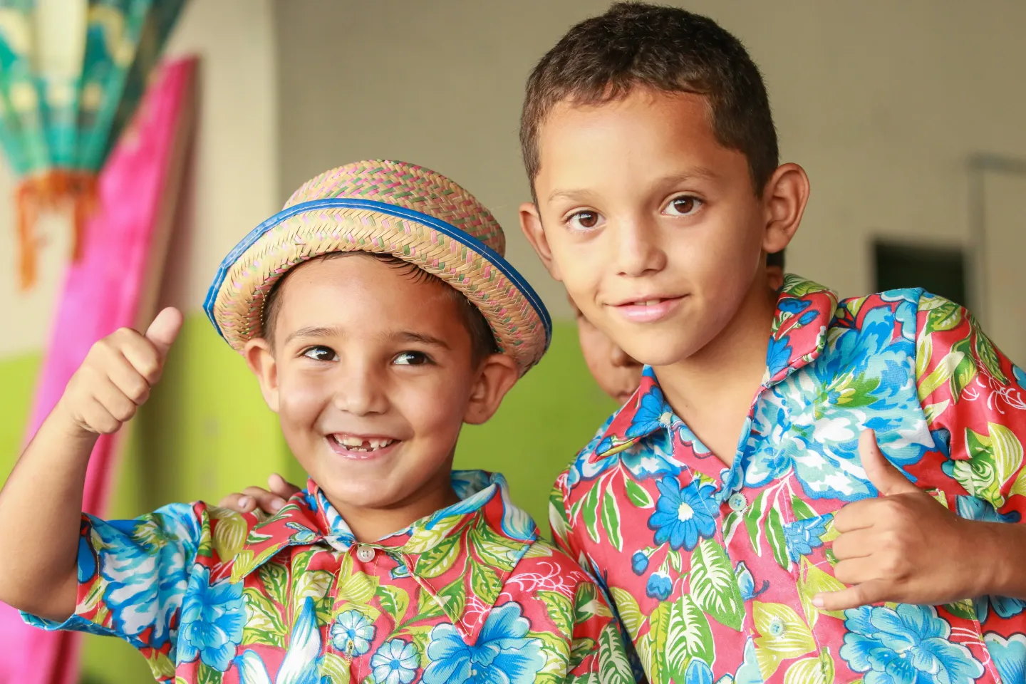 A foto mostra dois meninos com camisas floridas est&atilde;o de frente para a c&acirc;mera. O menor, &agrave; esquerda, est&aacute; olhando para o lado. Ele usa chap&eacute;u de palha. O outro, um pouco maior, est&aacute; olhando para a c&acirc;mera. Os dois est&atilde;o fazendo sinal de positivo com os dedos.