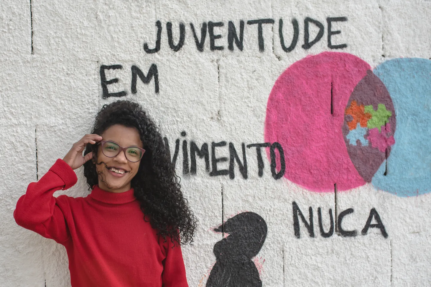 Foto mostra uma jovem sorridente, de &oacute;culos, parada em frente a um muro onde se l&ecirc; Juventude em Movimento Nuca. A jovem ajeita os cabelos compridos e cacheados, que est&atilde;o voando em seu rosto.