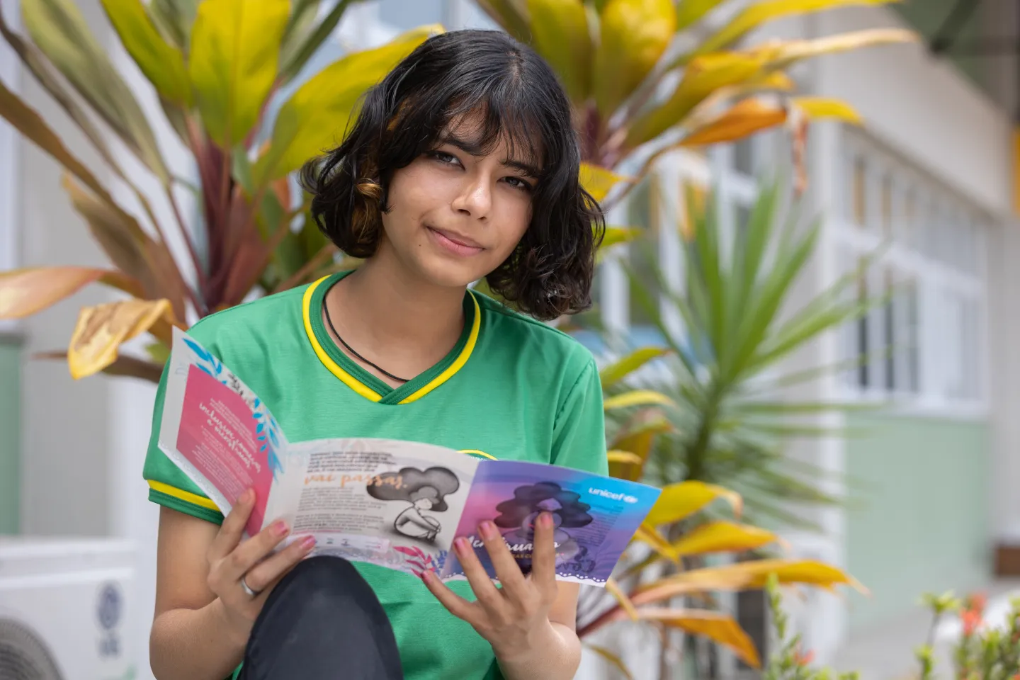 Foto mostra uma adolescente segurando um folder. Ela est&aacute; de uniforme escolar e olha para a c&acirc;mera.