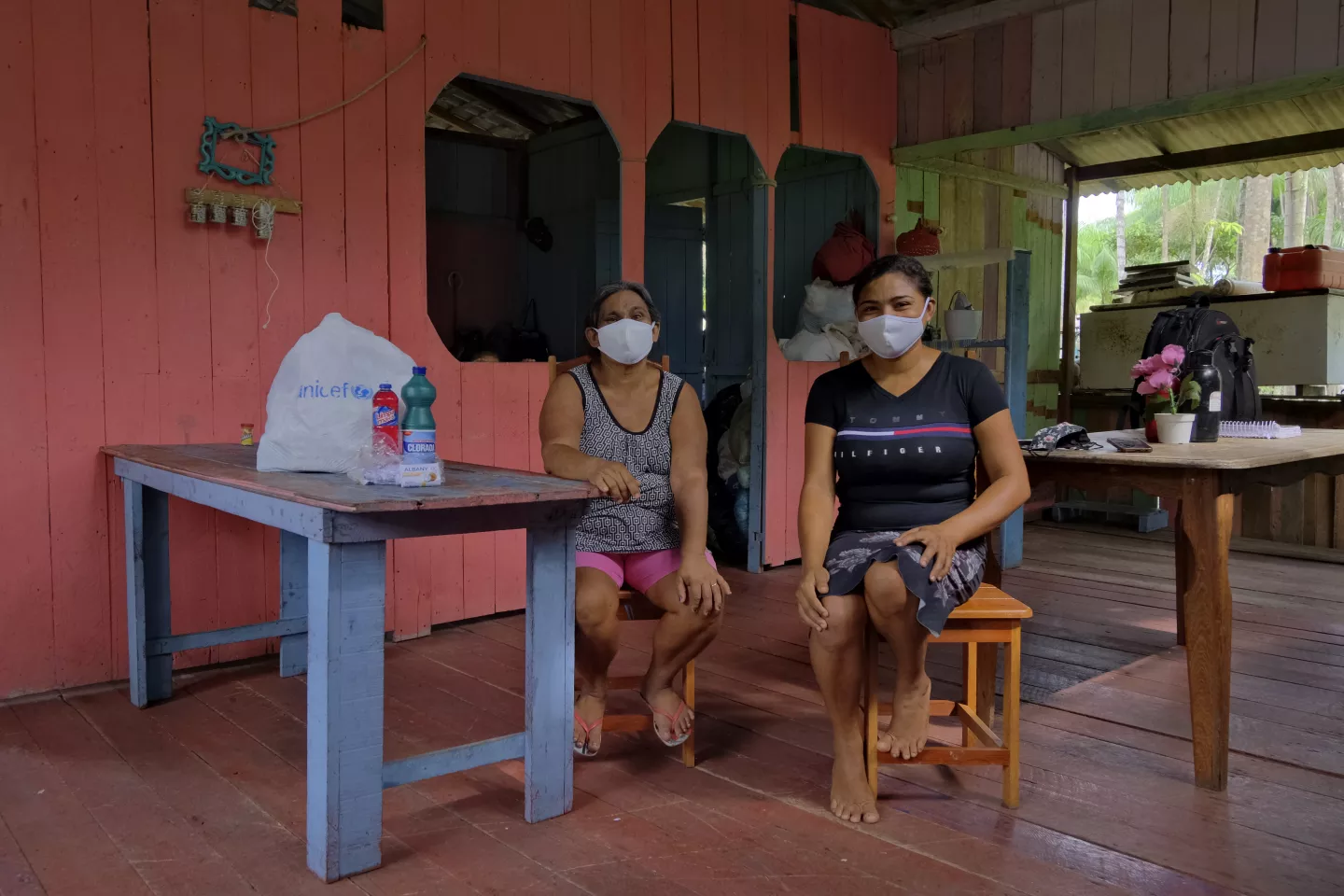 Duas mulheres, Lica e Joelma, est&atilde;o sentadas em um alpendre, ao lado de uma mesa com produtos doados pelo UNICEF.