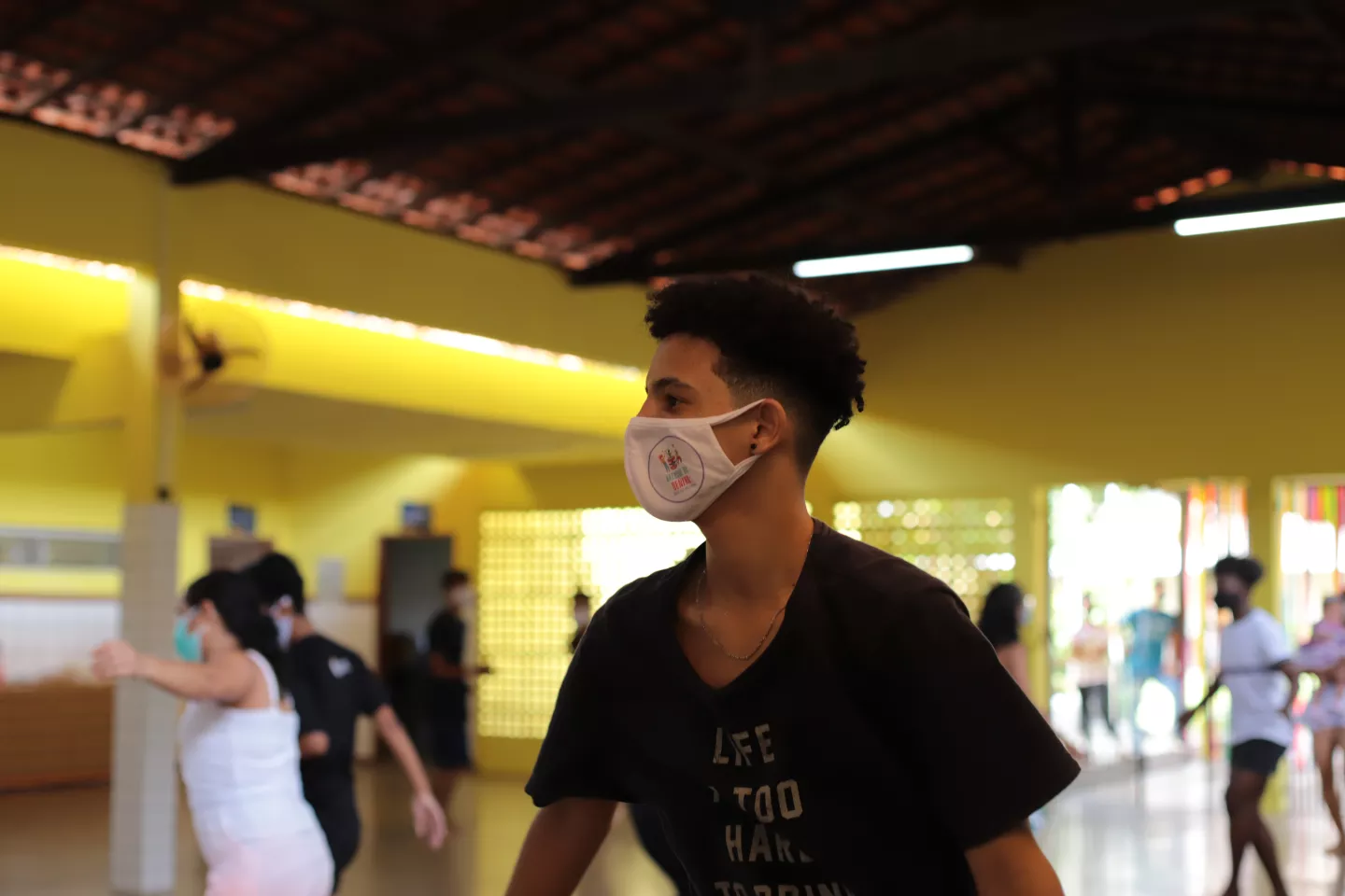 Foto mostra um adolescente dan&ccedil;ando em um sal&atilde;o bem espa&ccedil;oso. Ele usa m&aacute;scara. Ao fundo e ao redor, bem distantes umas das outras, h&aacute; outras pessoas dan&ccedil;ando tamb&eacute;m. Todos usam m&aacute;scara.