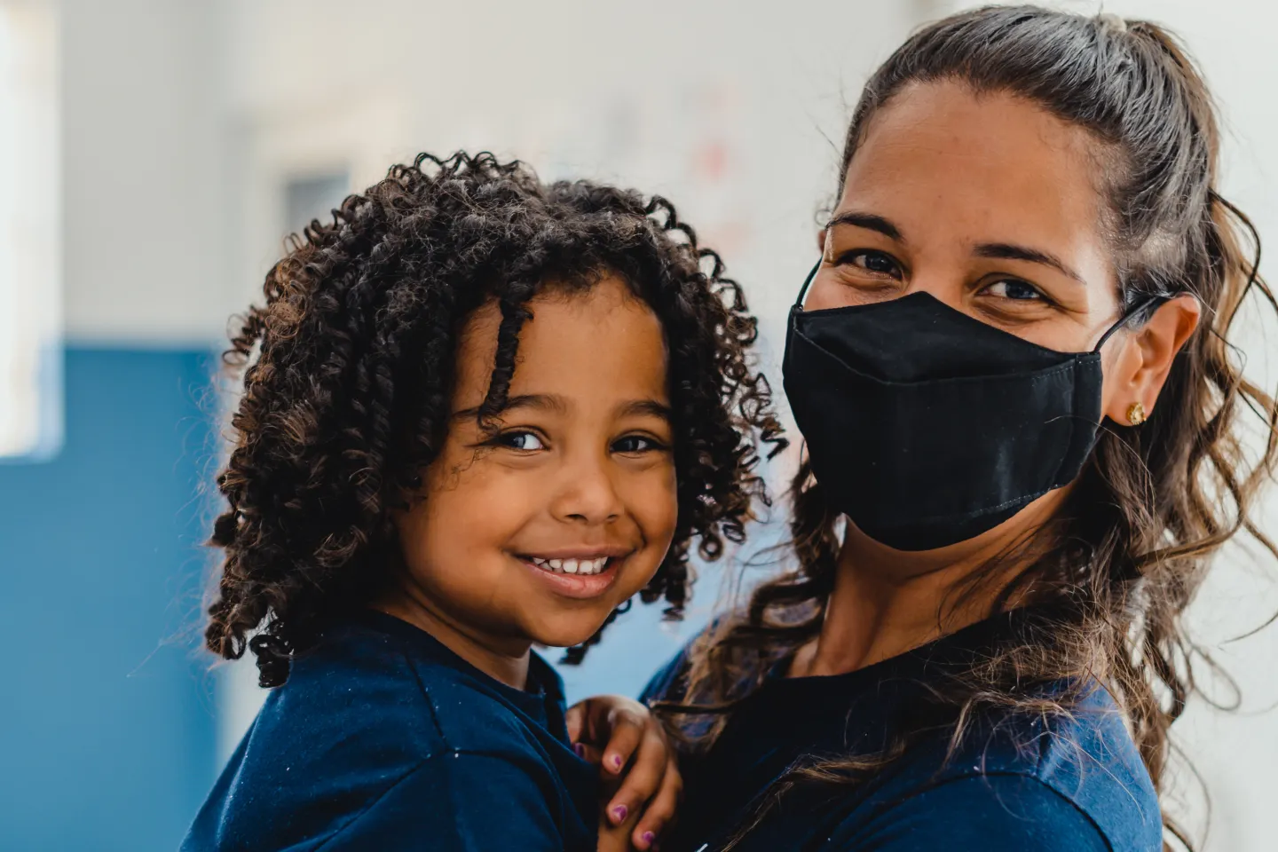 Foto mostra uma m&atilde;e usando m&aacute;scara segurando o filho pequeno no colo. Os dois est&atilde;o sorrindo.