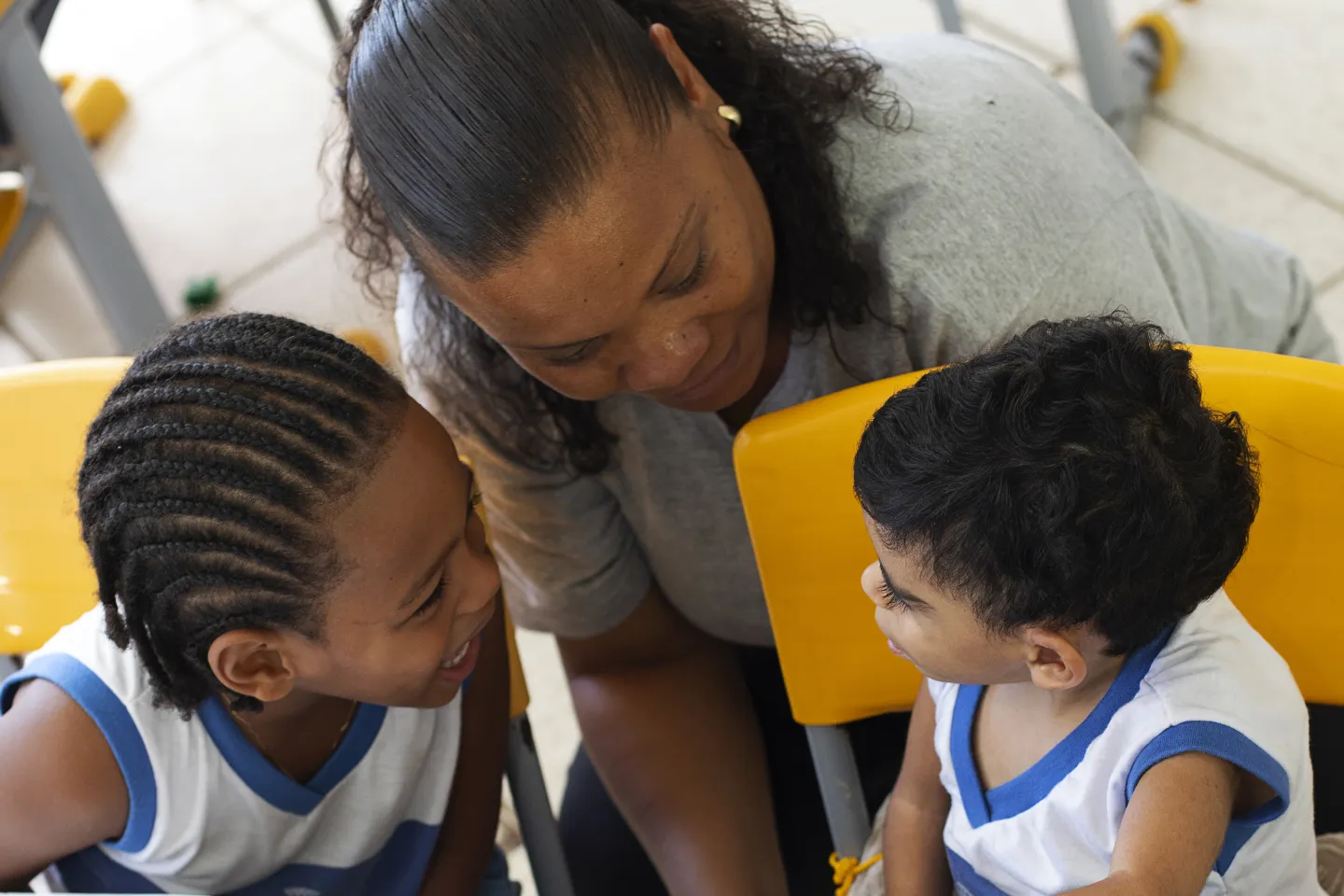 Foto mostra uma menina e um menino sentados na cadeira escolar ao lado de uma professora